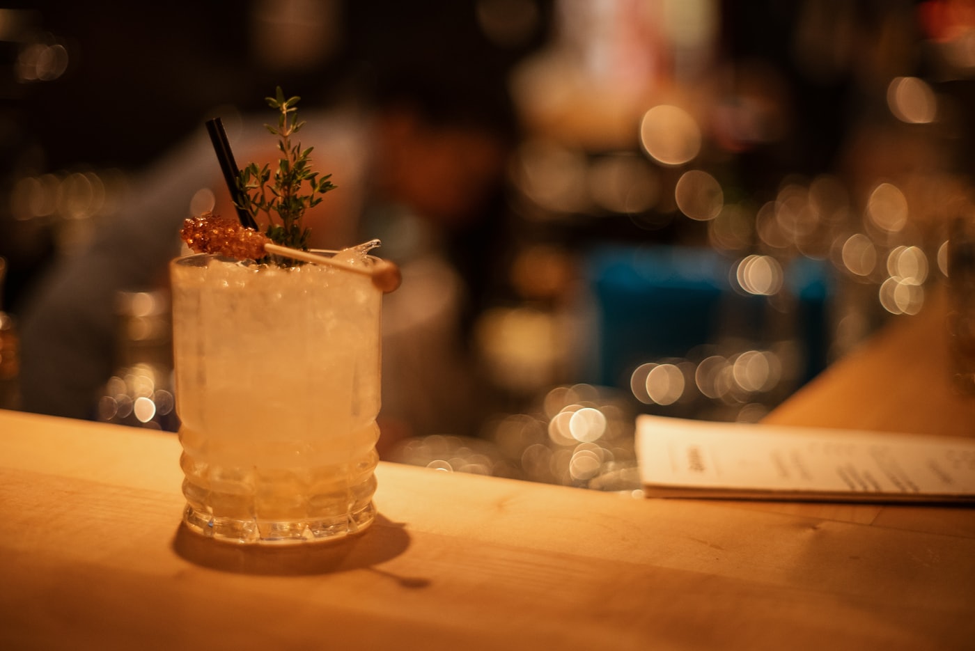 Cocktailglas mit Kräuterzweig auf einer Bar-Theke, warmes Licht und unscharfe Lichter im Hintergrund